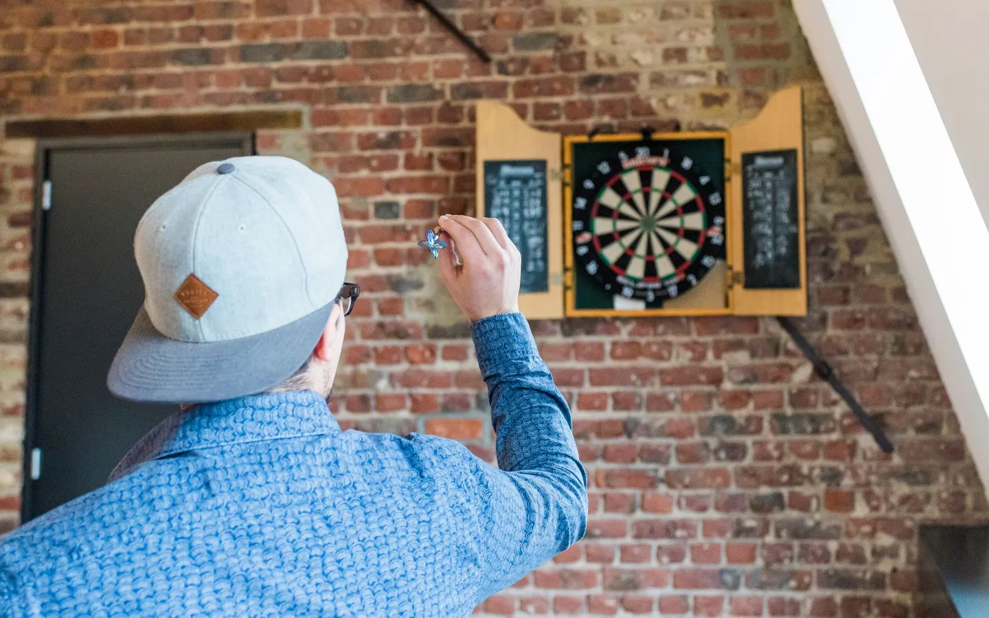 An adult in a blue shirt aiming a dart toward a dartboard in a brick-walled room